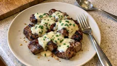 Stek wołowy z sosem serowym i szczypiorkiem na białym talerzu, widelcem i łyżką.