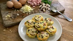 Omlety muffiny z grzybami, szynką i serem na talerzu, składniki na desce w tle.