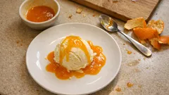 Lody waniliowe z sosem pomarańczowym na białym talerzu, obok miseczka i łyżka.