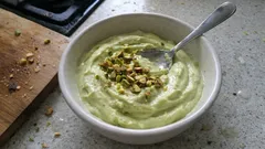 Pistacjowy deser w miseczce, posypany orzechami, ze srebrną łyżeczką, na blacie kuchennym.