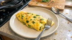 Omlet z ziołami na białym talerzu, na stole, z widelcem i łyżką obok.