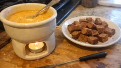 Fondue serowe w ceramicznym naczyniu z lampką i talerz grzanek na drewnianym stole.