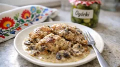 Kurczak w sosie pieczarkowym na talerzu, obok słoik ogórków kiszonych, dekoracyjna ściereczka.