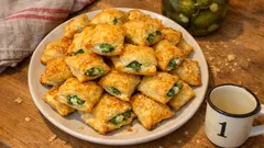 Talerzy pełen złocistych pasztecików ze szpinakiem na drewnianym stole, obok kubek i ściereczka.