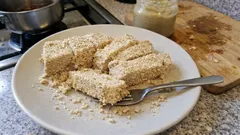 Talerz z pokrojoną chałwą sezamową na kuchennym blacie, widelec, słoik tahini w tle.