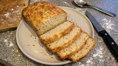 Domowy chleb pokrojony w plastry na talerzu, nóż i łyżka na blacie kuchennym.