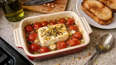 Pieczony ser feta z pomidorami i czosnkiem w naczyniu, grzanki na talerzu obok.