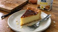 Kawałek sernika waniliowego na talerzu z widelcem, w tle składniki do pieczenia.