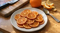Plasterki suszonej pomarańczy na talerzu, świeża pomarańcza i nóż na drewnianej desce.