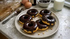 Domowe pączki z czekoladą na talerzu, obok szklanka mleka i składniki do pieczenia.