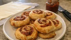 Cynamonowe ślimaczki na talerzu, obok słoik z dżemem i nóż na blacie kuchennym.