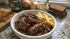 Gulasz wołowy z frytkami w misce, w tle chleb, piwo i cebula na kuchennym blacie.