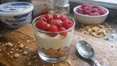 Deser z mascarpone, malinami i kruszonymi herbatnikami w szklance na drewnianej desce.